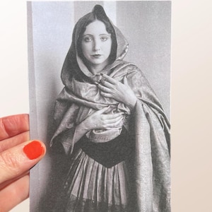 May include: A monochrome vintage photograph of a woman in a hooded cloak and dress. The woman is looking directly at the camera, hands clasped near her chest. The photograph has a soft focus and a vintage aesthetic, reminiscent of early photography.