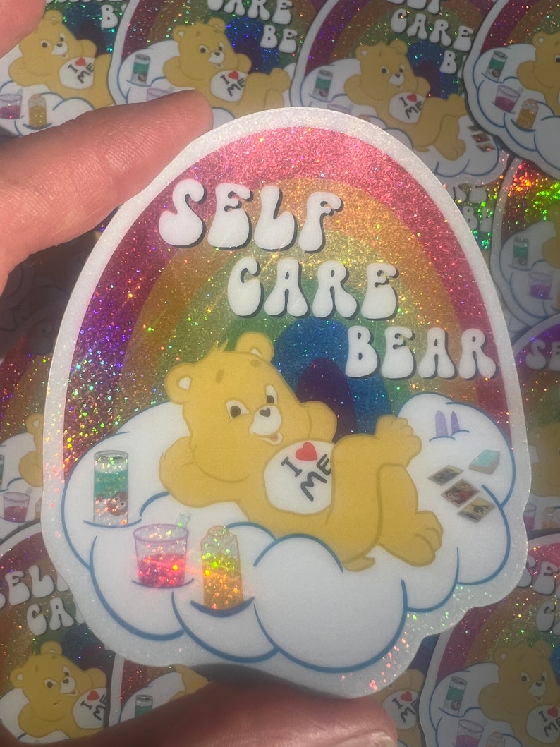 May include: A rainbow glitter sticker featuring a Care Bear with the text "SELF CARE BEAR" and "I &hearts; ME" on its belly. The bear is lying on a cloud with a rainbow background.