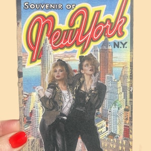 May include: A vintage postcard featuring two women in 1980s attire posing in front of a New York City skyline. The postcard reads "Souvenir of New York" in a retro font, with a blue sky background.