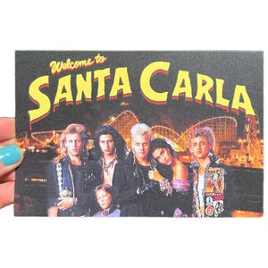 May include: A rectangular card featuring a group of people with the text "Welcome to SANTA CARLA" in yellow and red. The background shows a nighttime amusement park scene with a roller coaster.