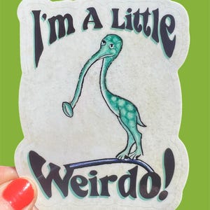 Peut inclure: Un autocollant avec une créature verte de dessin animé avec un long cou et un long nez. La créature se tient sur une ligne noire et le texte "I'm a little Weirdo!" est écrit en lettres noires.