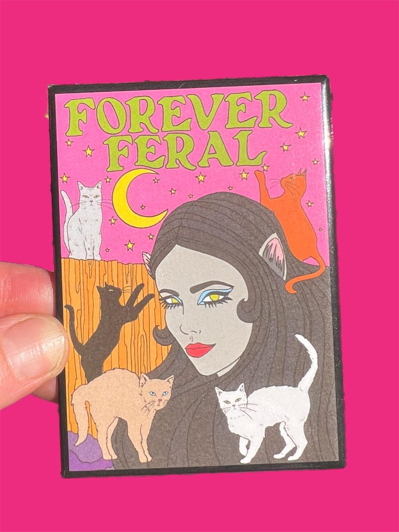 May include: A pink and green illustrated patch with the text "FOREVER FERAL" and a woman with cat ears surrounded by five cats.