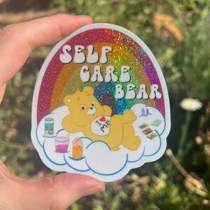 May include: A rainbow glitter sticker featuring a Care Bear character on a cloud. The bear is wearing a shirt that says "I love me". The sticker reads "Self Care Bear".