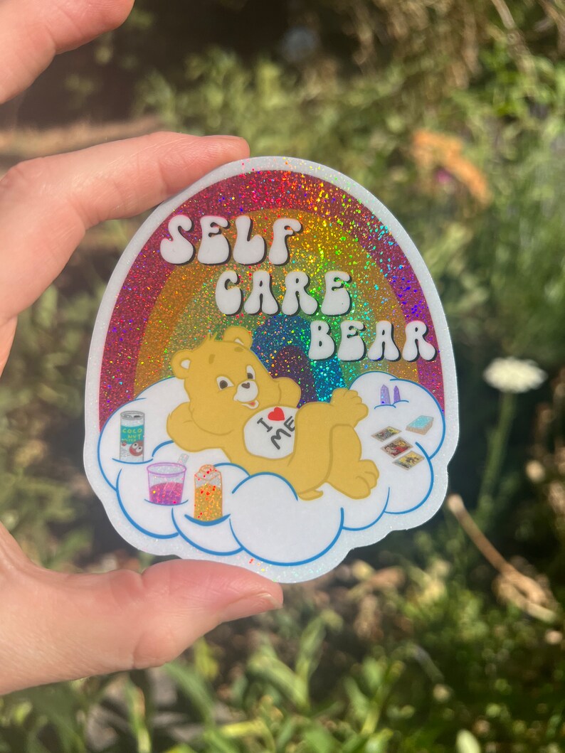 May include: A rainbow glitter sticker featuring a Care Bear character on a cloud. The bear is wearing a shirt that says "I love me". The sticker reads "Self Care Bear".