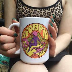 May include: White ceramic mug with a colorful graphic of a woman with a scorpion. The graphic says "Scorpio" in a retro font.