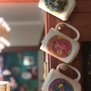May include: Three white ceramic mugs with colorful zodiac signs and symbols. The mugs are hanging on a wooden surface. The signs are Capricorn, Aries, and Aquarius.