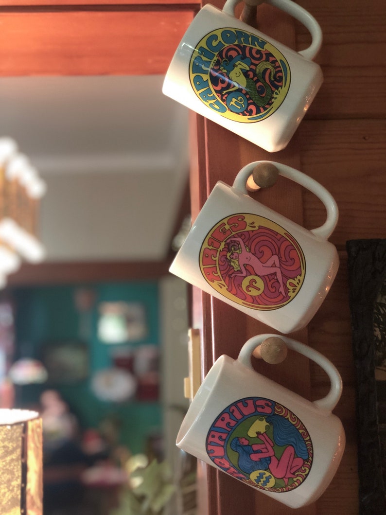 May include: Three white ceramic mugs with colorful zodiac signs and symbols. The mugs are hanging on a wooden surface. The signs are Capricorn, Aries, and Aquarius.
