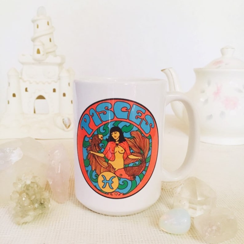 May include: White ceramic mug with a colorful graphic of a mermaid with the word "Pisces" in a circle around her. The mermaid is wearing a red cape and has long black hair.