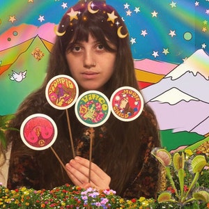 K&ouml;nnte beinhalten: Eine Frau mit langen braunen Haaren h&auml;lt vier runde Aufkleber auf St&ouml;cken. Die Aufkleber zeigen astrologische Zeichen: Skorpion, Jungfrau, Mondkind. Der Hintergrund ist eine farbenfrohe psychedelische Landschaft mit Bergen, Sternen und V&ouml;geln.