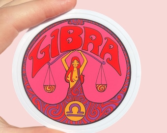 May include: A pink and purple circle sticker with the word "LIBRA" in red letters. A woman with red hair is holding a scale with two bowls. The scales are surrounded by a purple and red swirl design. The Libra symbol is in the center of the sticker.