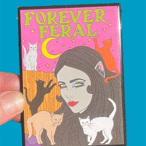 May include: A pink and black patch with the text "FOREVER FERAL" in green letters. The patch features a woman with long black hair and cat ears, surrounded by five cats. The cats are white, orange, black, and brown.