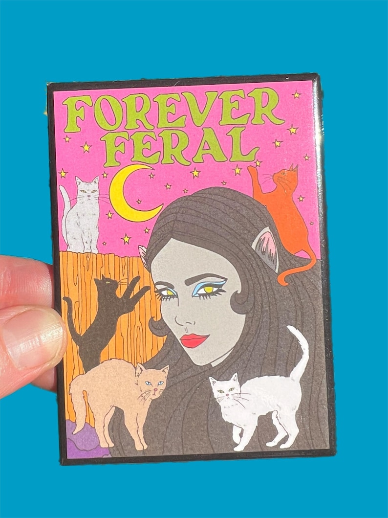 May include: A pink and black patch with the text "FOREVER FERAL" in green letters. The patch features a woman with long black hair and cat ears, surrounded by five cats. The cats are white, orange, black, and brown.