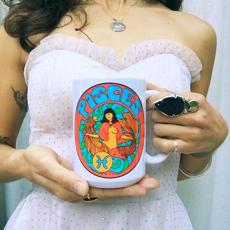 May include: White ceramic mug with a colorful graphic of a woman with a fish tail and the word "Pisces" in a retro font.