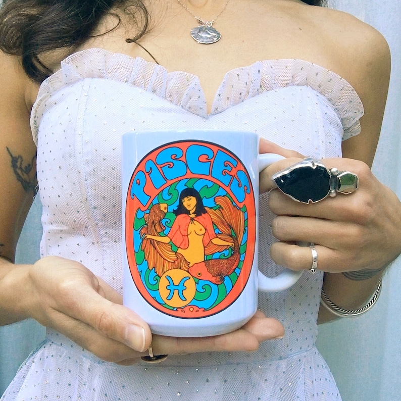 May include: A white ceramic mug with a colorful illustration of a mermaid with the word "Pisces" in a retro font. The mermaid is surrounded by fish and seaweed.
