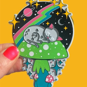May include: A white cat with black spots sleeps on a green mushroom with white polka dots. The mushroom is surrounded by stars and a rainbow. The background is black with a pink planet and a crescent moon.