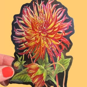 May include: A sticker featuring a vibrant dahlia flower with red and yellow petals, green leaves, and a black background. The flower has glitter accents, adding a sparkling effect. The sticker is held by a hand with a red painted nail.