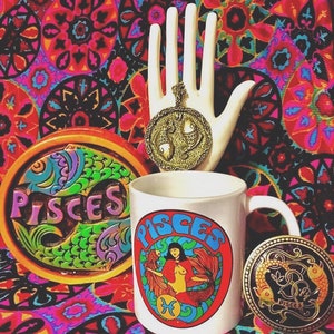 May include: A white ceramic mug with a colorful Pisces zodiac sign graphic. The mug has a black handle and is surrounded by a colorful patterned background. There is also a gold-colored Pisces zodiac pendant and a coaster with a Pisces zodiac sign.