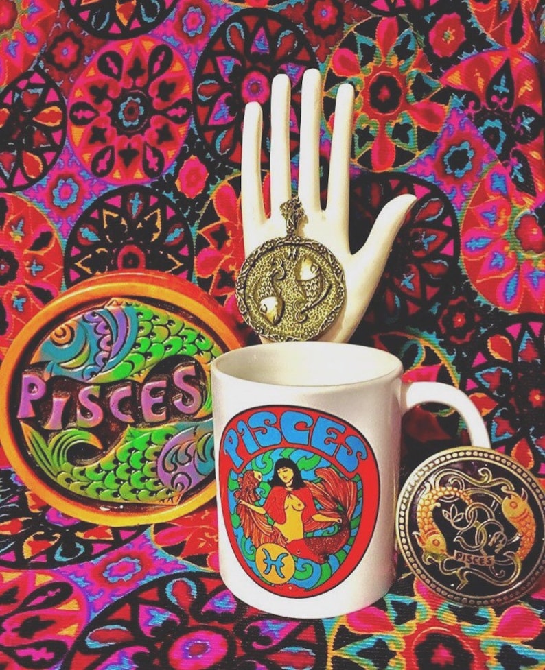 May include: A white ceramic mug with a colorful Pisces zodiac sign graphic. The mug has a black handle and is surrounded by a colorful patterned background. There is also a gold-colored Pisces zodiac pendant and a coaster with a Pisces zodiac sign.
