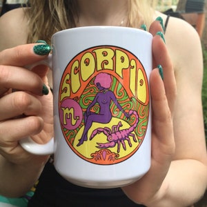 May include: White ceramic mug with a colorful graphic of a woman sitting on a scorpion with the word "Scorpio" in a psychedelic font.
