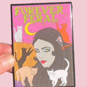 May include: A pink and green illustrated pin with the text "FOREVER FERAL" and a woman with cat ears surrounded by cats.