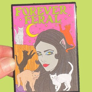 May include: A pink and black enamel pin with the text "FOREVER FERAL" in green letters. The pin features a woman with cat ears and a crescent moon behind her. There are five cats surrounding her, including a white cat, a black cat, a brown cat, and two orange cats.