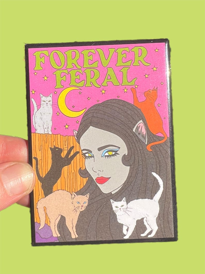 May include: A pink and black enamel pin with the text "FOREVER FERAL" in green letters. The pin features a woman with cat ears and a crescent moon behind her. There are five cats surrounding her, including a white cat, a black cat, a brown cat, and two orange cats.