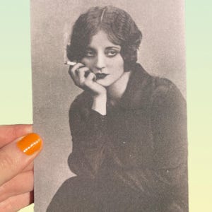 May include: A black and white vintage photograph of a woman with dark hair, dark lipstick, and a cigarette. The woman is resting her chin on her hand, with a contemplative expression. The photograph is held by a hand with orange nail polish.