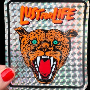Peut inclure: Autocollant avec un fond holographique et une bordure noire. L'autocollant présente une tête de léopard orange avec des yeux verts et le texte "LUST FOR LIFE" en orange. La gueule du léopard est ouverte, révélant un intérieur rouge et des dents blanches.
