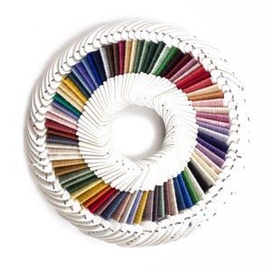 May include: A circular arrangement of numerous embroidery floss bobbins, each wrapped with a different solid color thread. The white cardboard bobbins are fan-shaped and feature small printed text, displaying a wide spectrum of vibrant and neutral hues.