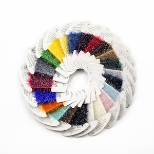 May include: A circular arrangement of numerous small, white cardboard cards, each holding a sample of Wispy Fringe yarn in a different color. The yarn samples feature a metallic, shimmering texture and are displayed in a vibrant spectrum of hues.