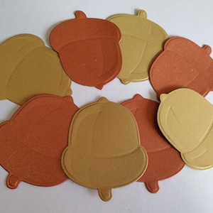 May include: A collection of die-cut paper acorns in shades of gold and rust orange. The acorn shapes are arranged in a circular pattern, with varying textures and tones, suggesting a fall theme.