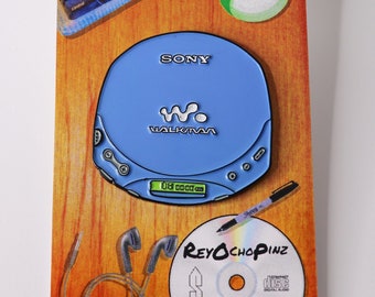 May include: A blue Sony Walkman CD player enamel pin with the text "SONY" and "WALKMAN" on the front. The pin is on a brown wooden background with a CD, a pen, and headphones.