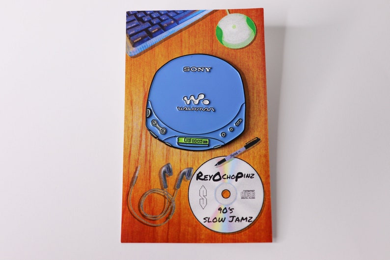 May include: A blue Sony Walkman CD player enamel pin with the text "SONY" and "WALKMAN" on the front. The pin is on a brown wooden background with a CD, a pen, and headphones.