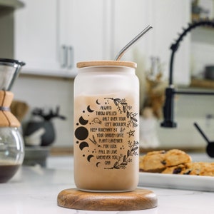 May include: A frosted glass tumbler with a wooden lid and a stainless steel straw. The tumbler has a black and white design with a moon phase pattern, stars, and the text "ALWAYS THROW SPILLED SALT OVER YOUR LEFT SHOULDER KEEP ROSEMARY BY YOUR GARDEN GATE PLANT LAVENDER FOR LUCK FALL IN LOVE WHENEVER YOU CAN".