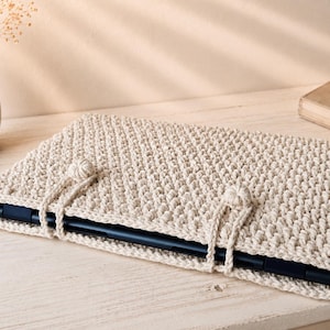 Könnte beinhalten: Eine beige gehäkelte Laptoptasche mit einem strukturierten Stichmuster. Die Tasche hat einen Klappenverschluss mit einem geknoteten Detail. Ein schwarzer Laptop ist teilweise sichtbar. Die Tasche liegt auf einer hellen Holzoberfläche.