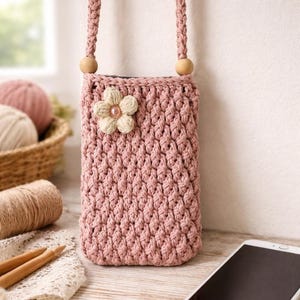 May include: A dusky pink crochet phone case with a braided strap and wooden beads. A cream flower with a pearl centre decorates the front. The case is next to a smartphone and craft supplies.