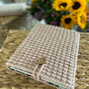 Puede incluir: Una funda de libro de crochet rosa con cierre de botón marrón. La funda está sobre una cesta tejida.