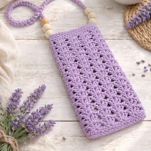 Puede incluir: Una funda de teléfono de ganchillo lila con una correa y cuentas de madera. La funda tiene un diseño calado y un lazo para llevar. El fondo incluye ramitas de lavanda y un objeto de cerámica blanca.