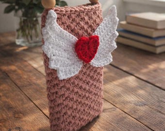 Puede incluir: Un bolso de ganchillo en color rosa empolvado con un corazón rojo central flanqueado por dos alas de ganchillo blancas. El bolso tiene un asa superior formada por dos grandes cuentas de madera y un tejido denso y texturizado.
