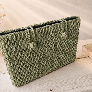 May include: A sage green rectangular crochet handbag featuring a textured diamond stitch pattern, two thin matching cord handles, and small decorative knot details at the base of each handle attachment point on the front exterior.