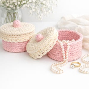 May include: Two handmade crochet jewelry boxes in cream and pink, one open with a pearl necklace inside. A gold ring with a diamond sits nearby. The boxes have lids with pink pom-pom accents. White flowers are in the background.