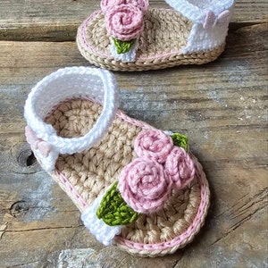 May include: A pair of crocheted baby sandals with pink and green flower embellishments. The sandals are made with a beige and white yarn.