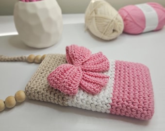 Puede incluir: Una funda de teléfono de ganchillo hecha a mano con un lazo rosa, secciones beige, blancas y rosas, y una correa de cuentas de madera. La funda está diseñada con un patrón de puntada texturizada. Hay ovillos de lana en el fondo.