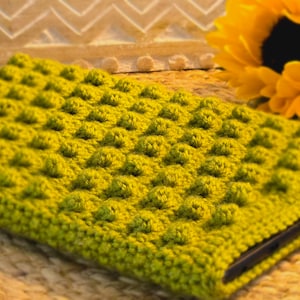 May include: A green crocheted phone case with a textured, bumpy surface. The case is lying on a woven straw mat.