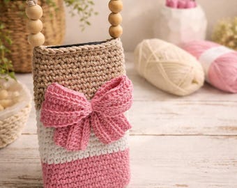 May include: Crocheted phone case in beige, white, and pink, featuring a large pink bow. The case has a braided strap with wooden beads. The background includes yarn and a basket.