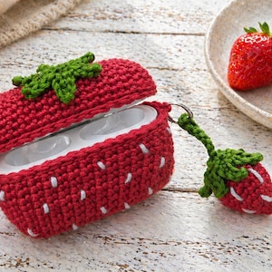 May include: A red crocheted AirPod case shaped like a strawberry, with white seed details and a green leaf top. A matching strawberry keychain is attached. Fresh strawberries sit on a white plate.