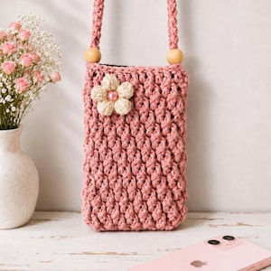 Puede incluir: Un bolso de teléfono de ganchillo rosa empolvado con una correa trenzada y cuentas de madera. El bolso presenta una flor color crema con un centro rosa. Un smartphone rosa descansa sobre una superficie de madera blanca.