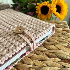 May include: A pink crocheted book cover with a wooden button closure. The cover is wrapped around a book with a floral design. The book is sitting on a woven basket.