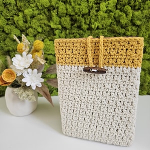 Op de afbeelding: Een gehaakte telefoonhoes met een mosterdgele bovenkant en een crèmekleurige onderkant. De hoes heeft een houten sluiting.