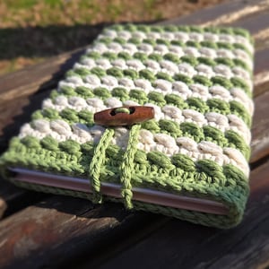 May include: A green and white crocheted book cover with a wooden button closure. The cover is made with a textured stitch pattern.